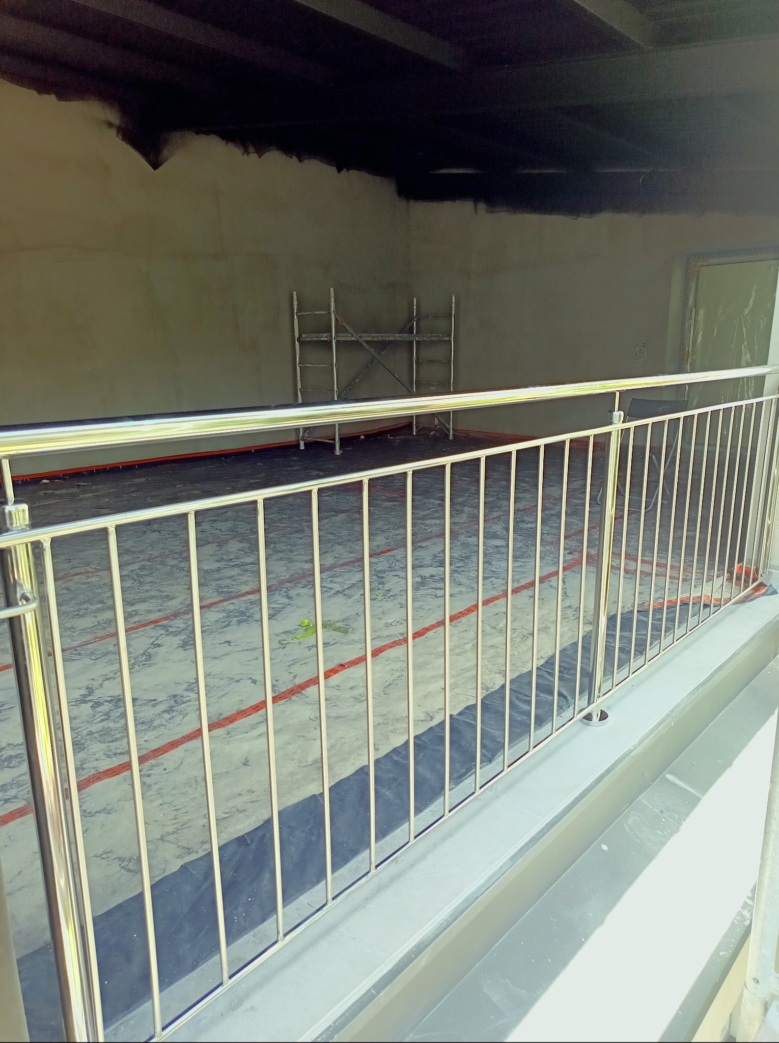 Barierka ze stali nierdzewnej na tle surowego balkonu w trakcie wykańczania. Widoczne rusztowanie i ślady farby na ścianach.