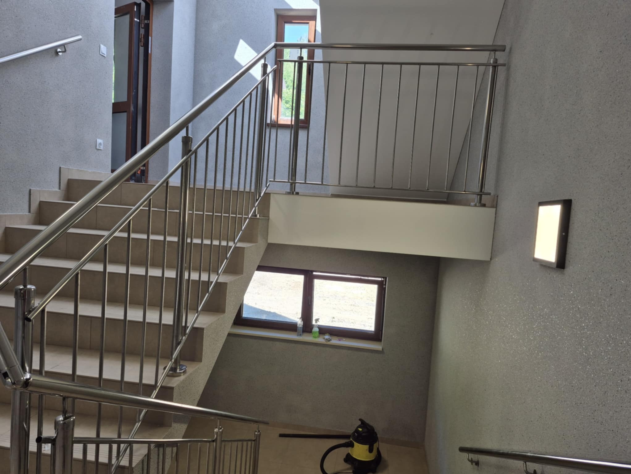 Wnętrze klatki schodowej z jasnymi schodami i metalową balustradą, widok na okna w brązowych ramach. Na podłodze widoczny odkurzacz.