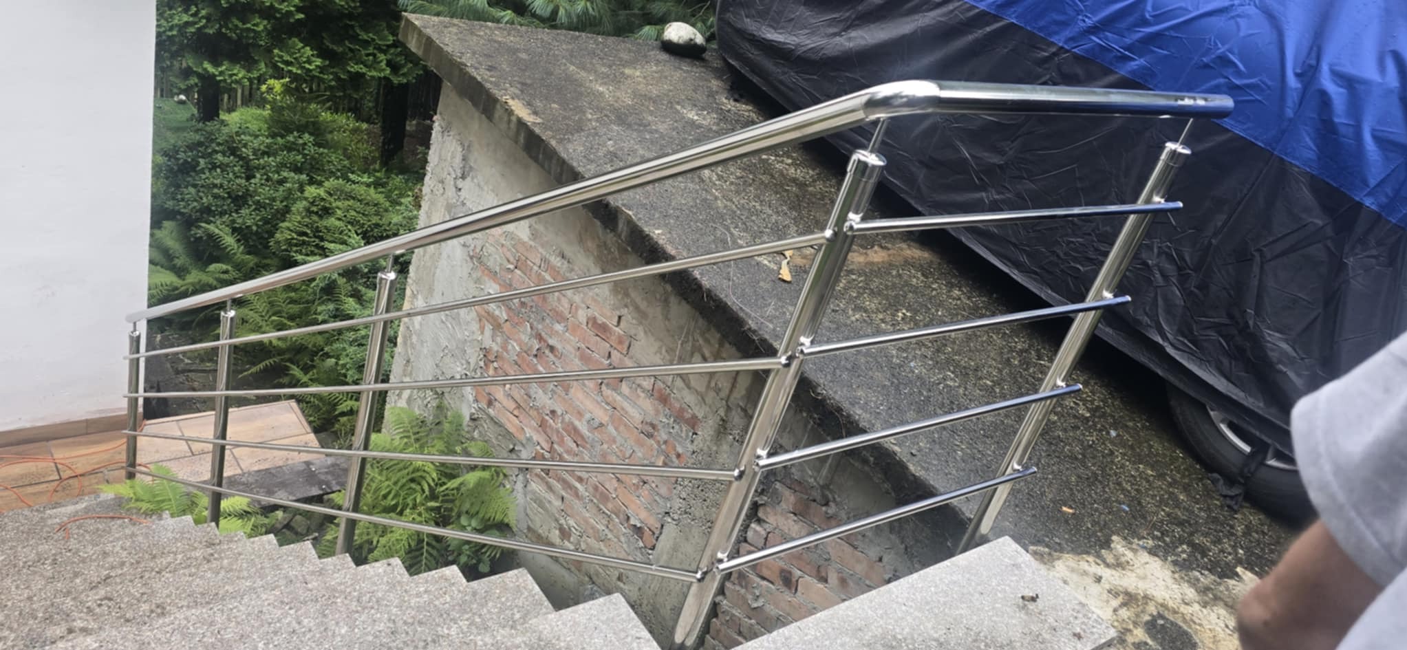 Polerowana balustrada ze stali nierdzewnej na schodach zewnętrznych, z widokiem na zieloną roślinność i fragment muru z cegły w tle.