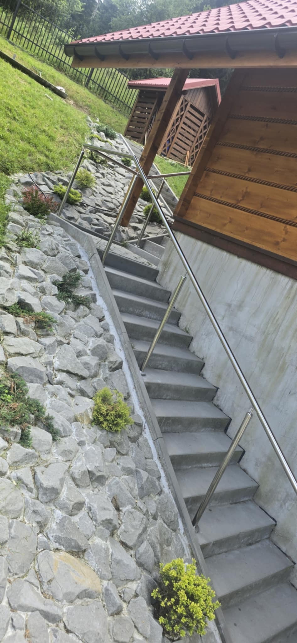 Betonowe schody z nierdzewną poręczą, wkomponowane w kamienny mur na zboczu, prowadzące do drewnianej altany w tle. Ujęcie z dołu, podkreślające strome nachylenie.