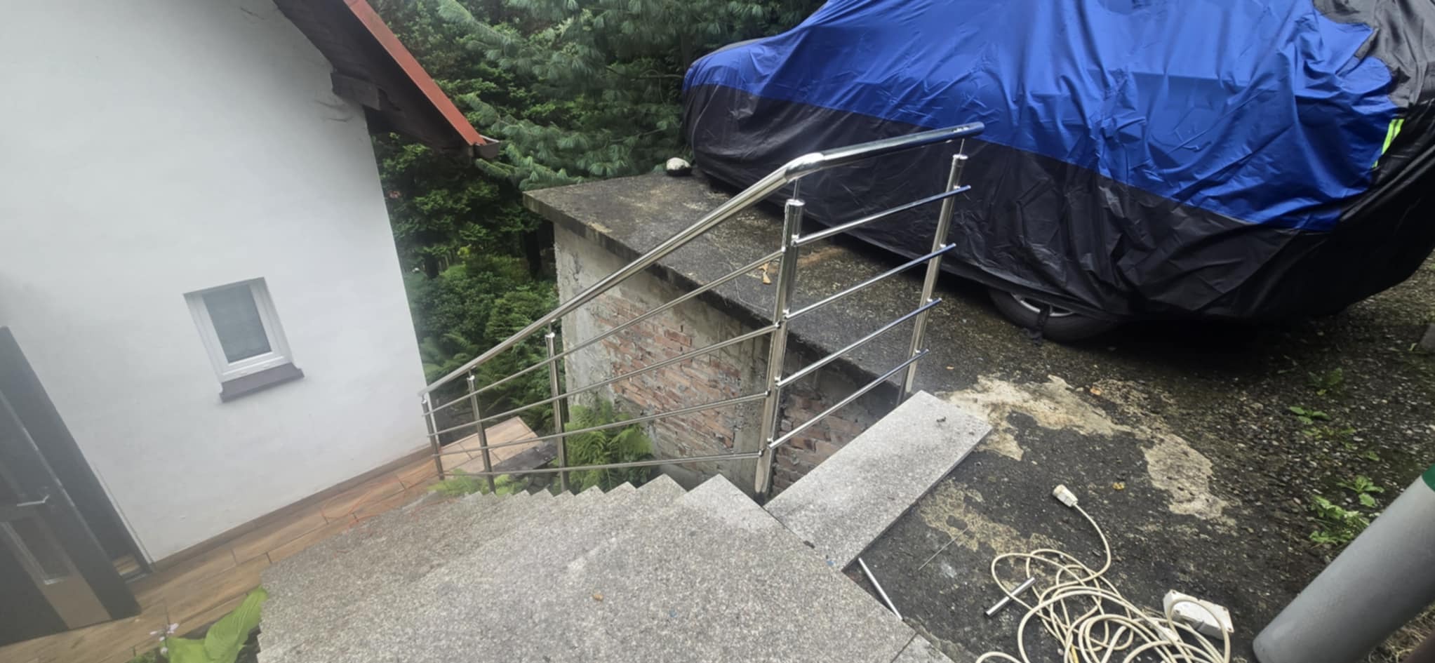 Stalowa balustrada schodowa z rurek, zamontowana na zewnątrz budynku w Żabnicy, z widocznym zadaszeniem na samochód w tle. Ujęcie z góry, schody betonowe.