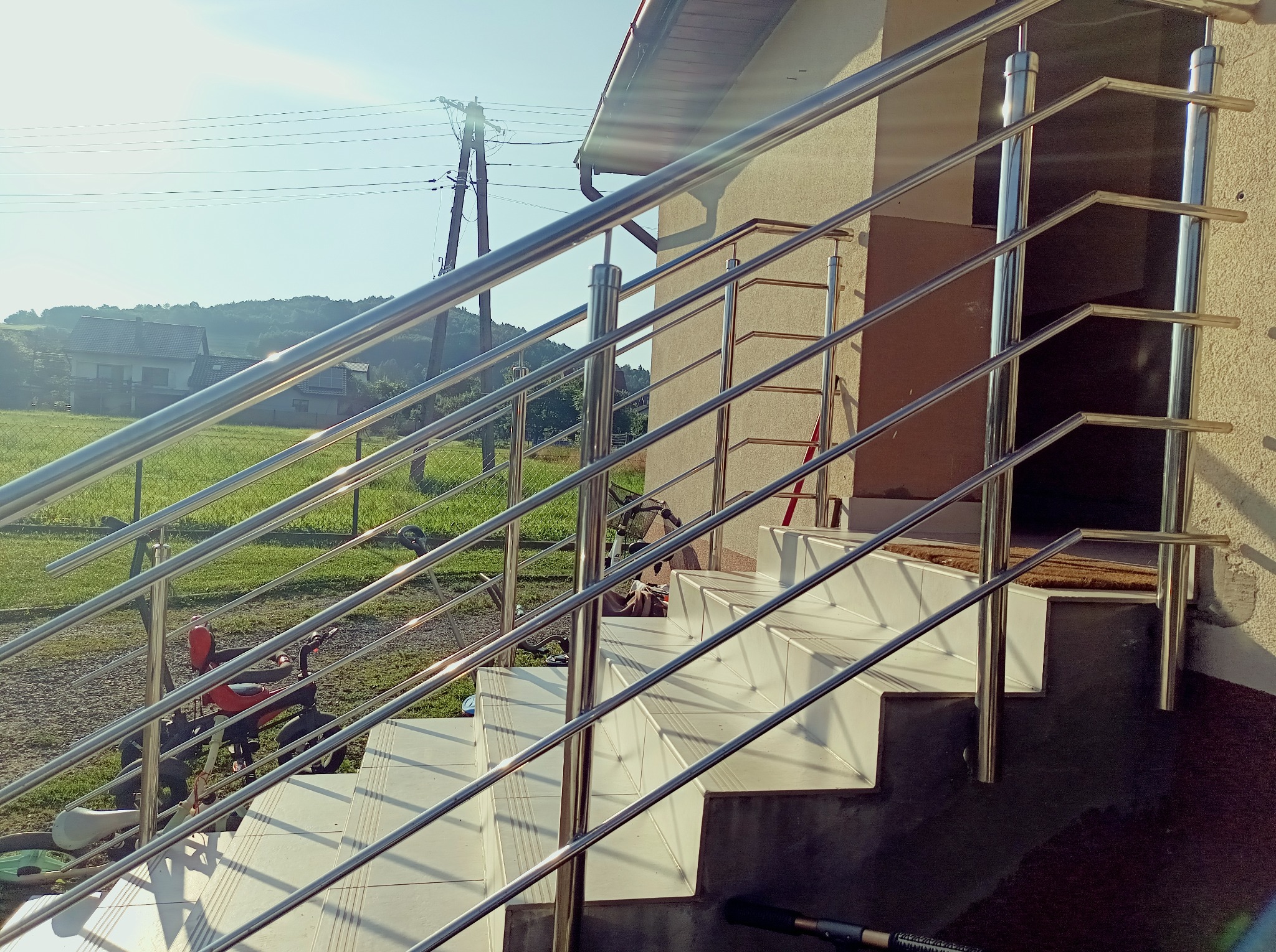 Zewnętrzne schody z jasnymi płytkami i lśniącą, metalową balustradą w słoneczny dzień. W tle dom z ogrodem i pagórkowaty krajobraz.