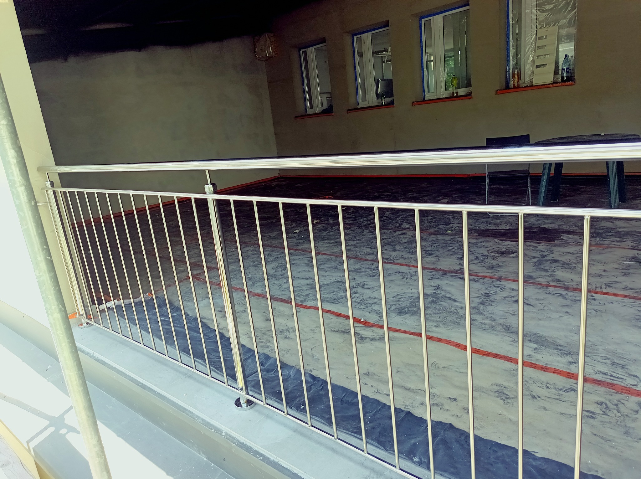 Błyszcząca, metalowa balustrada z pionowymi prętami na tle remontowanego pomieszczenia z oknami i stolikiem. Widoczne zabezpieczenia okien folią.