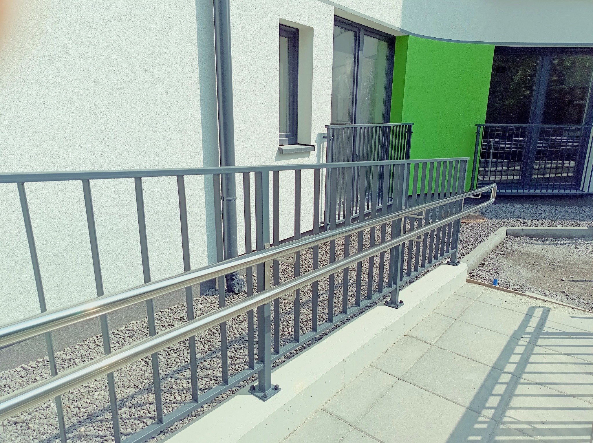 Szara, metalowa balustrada schodowa z poręczą ze stali nierdzewnej na zewnątrz budynku z białymi ścianami i zielonym akcentem. Widoczne płytki chodnikowe i żwir.