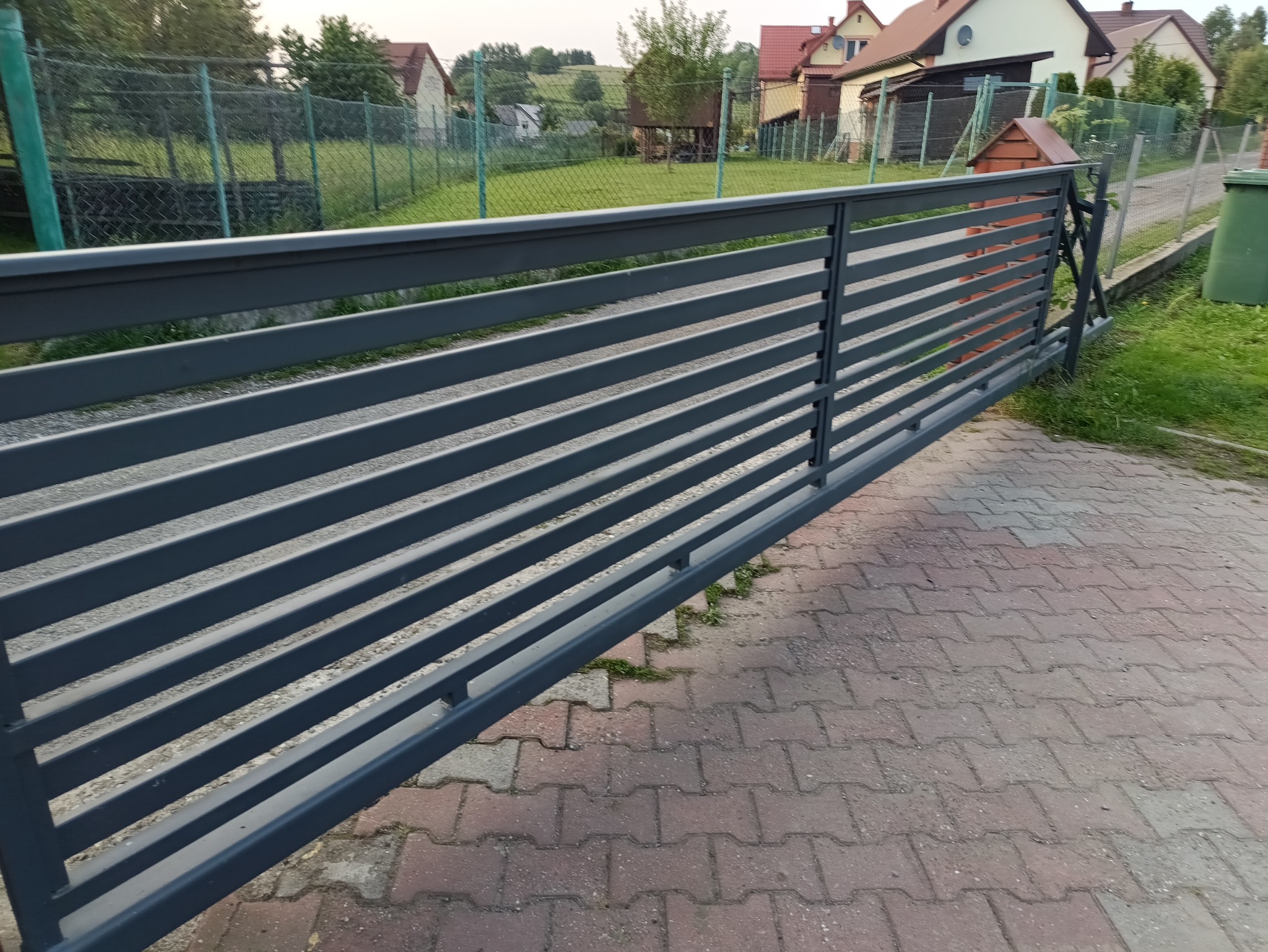 Nowoczesna, przesuwna brama posesyjna z poziomych, szarych profili. W tle zabudowa jednorodzinna i zieleń. Widoczne detale konstrukcyjne i wykończenie.