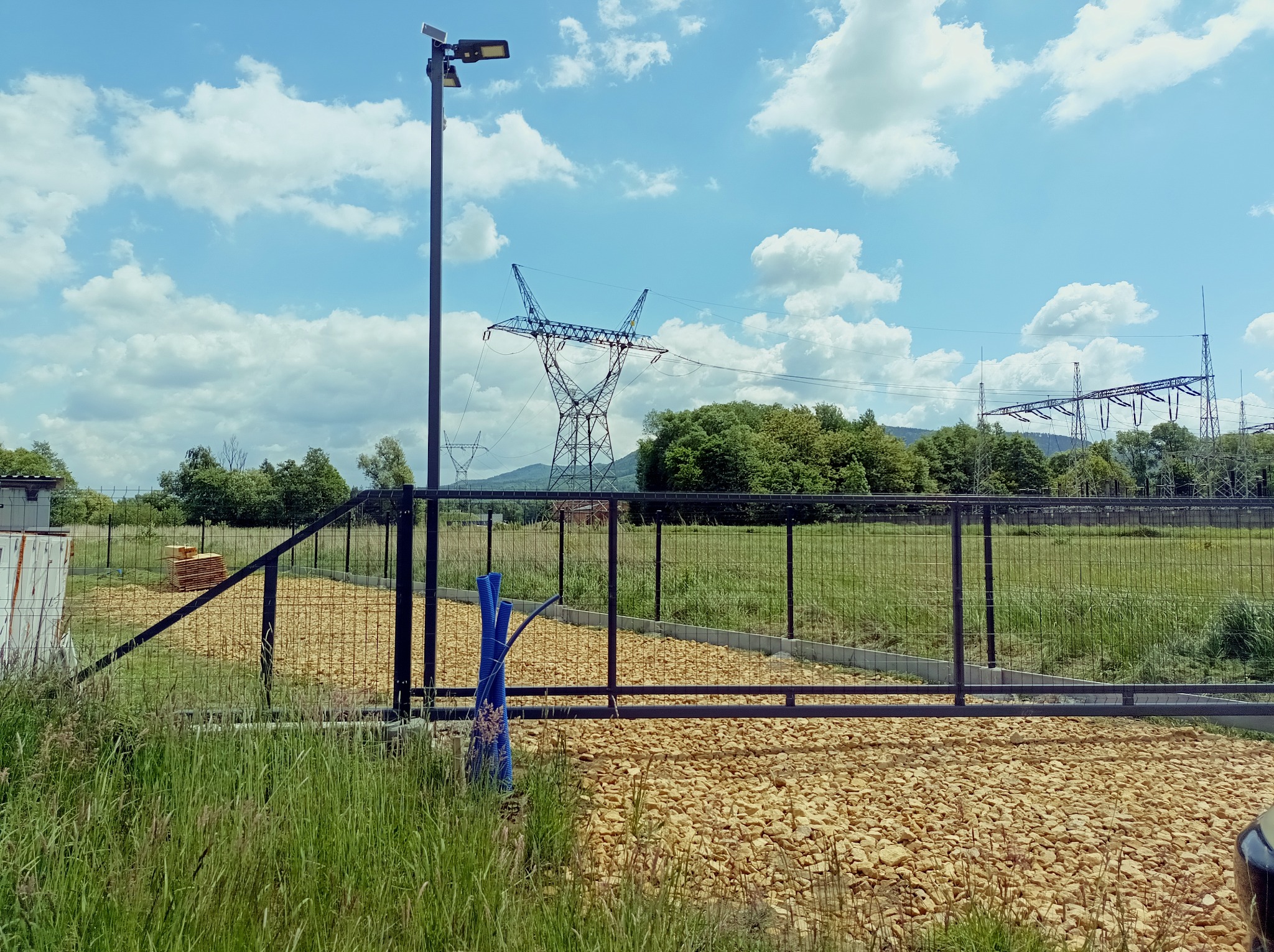 Metalowa brama wjazdowa z siatki na posesję, z widoczną infrastrukturą energetyczną w tle. Teren wysypany kamieniami.