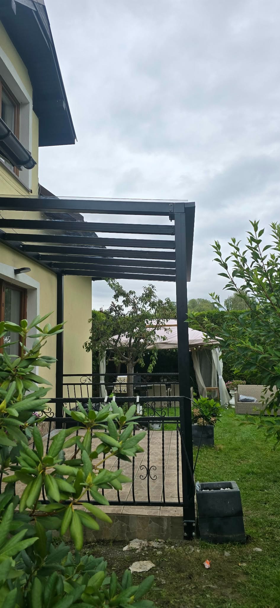 Czarna pergola metalowa z zadaszeniem, przylegająca do budynku. Widok z boku, z elementami ogrodu w tle i na pierwszym planie. Zastosowano ozdobne balustrady.