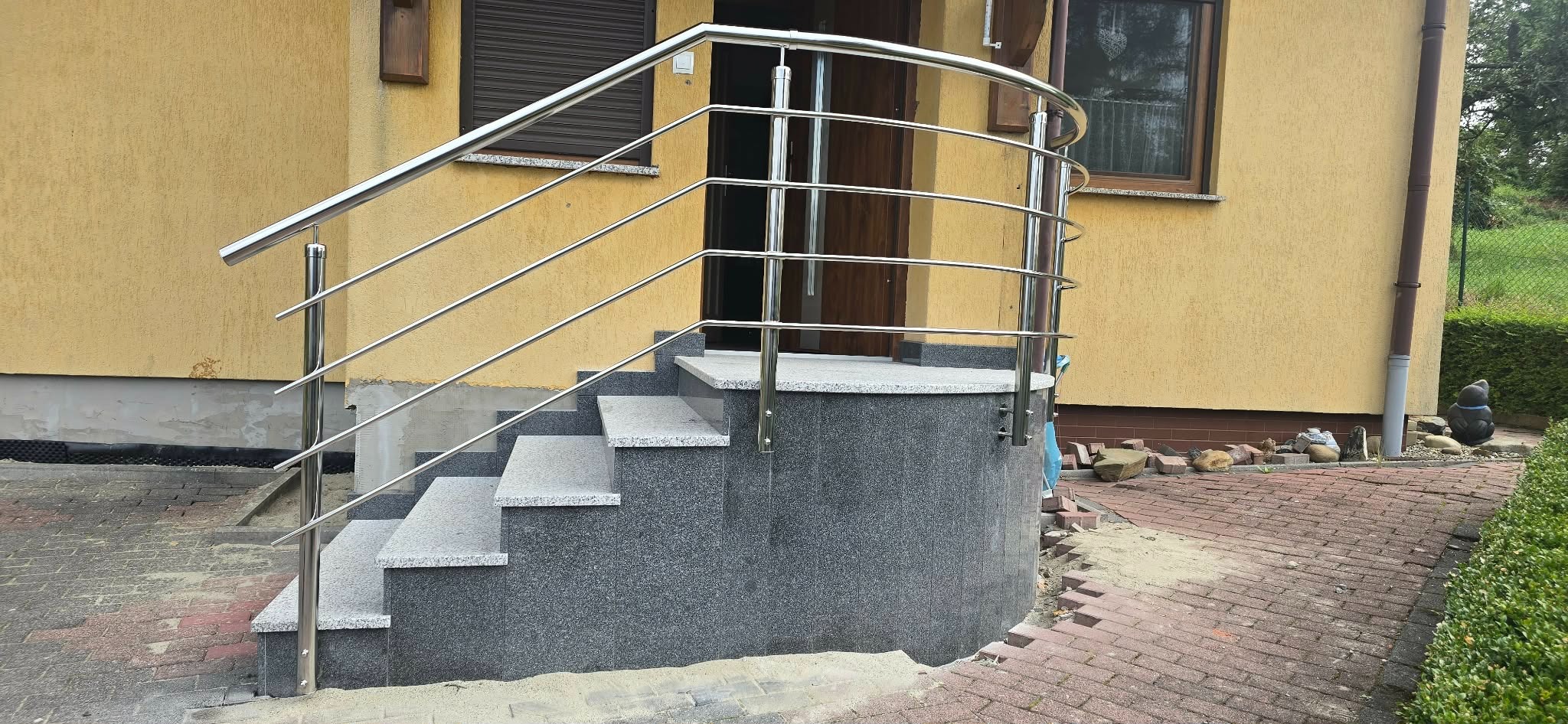 Zewnętrzne schody z szarego granitu z balustradą ze stali nierdzewnej, prowadzące do drzwi wejściowych domu o żółtej elewacji. Widoczne detale wykończenia i otoczenie.