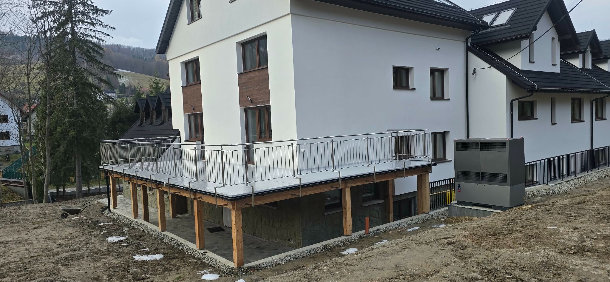 Nowoczesny balkon z balustradą ze stali nierdzewnej w Żabnicy. Jasna elewacja budynku, drewniane elementy konstrukcyjne balkonu. Widok z dołu, częściowo zasypany teren.