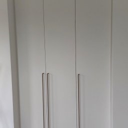 N&M Meble - Biała szafa wnękowa z trzema drzwiami i długimi, pionowymi uchwytami w kolorze brązu, ustawiona na drewnianej podłodze przy białej ścianie.