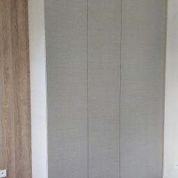 N&M Meble - Jasnoszara szafa wnękowa z trzema drzwiami, dopasowana do sufitu, na tle ściany z drewnianą okładziną i ciemnej podłogi. Widoczny fragment okrągłego blatu stołu.