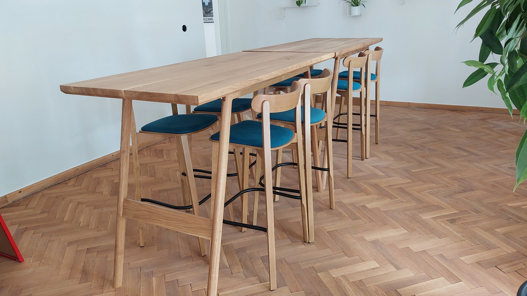 Długi, jasny drewniany stół barowy z sześcioma krzesłami barowymi z turkusowymi siedziskami na parkiecie w jodełkę, oświetlony naturalnym światłem.