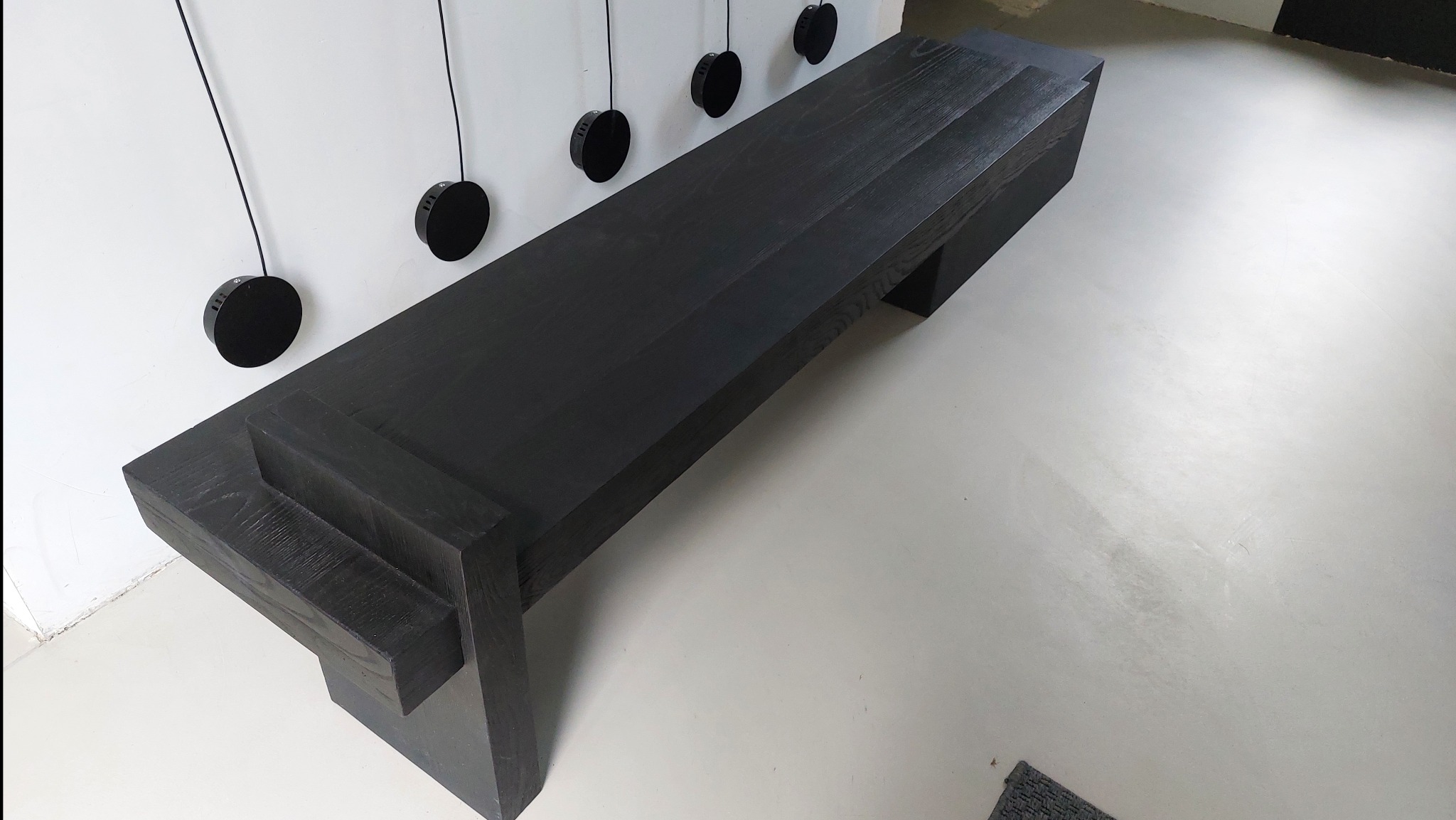 Długi, czarny, drewniany stół/ławka z wyraźnym usłojeniem drewna, minimalistyczny design, z czarnymi lampami na ścianie.