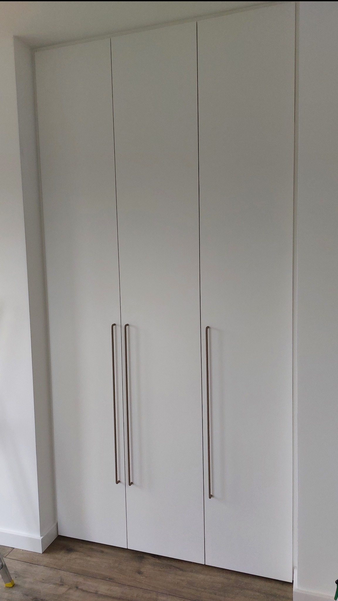 Biała szafa wnękowa z trzema drzwiami i długimi, pionowymi uchwytami w kolorze brązu, ustawiona na drewnianej podłodze przy białej ścianie.