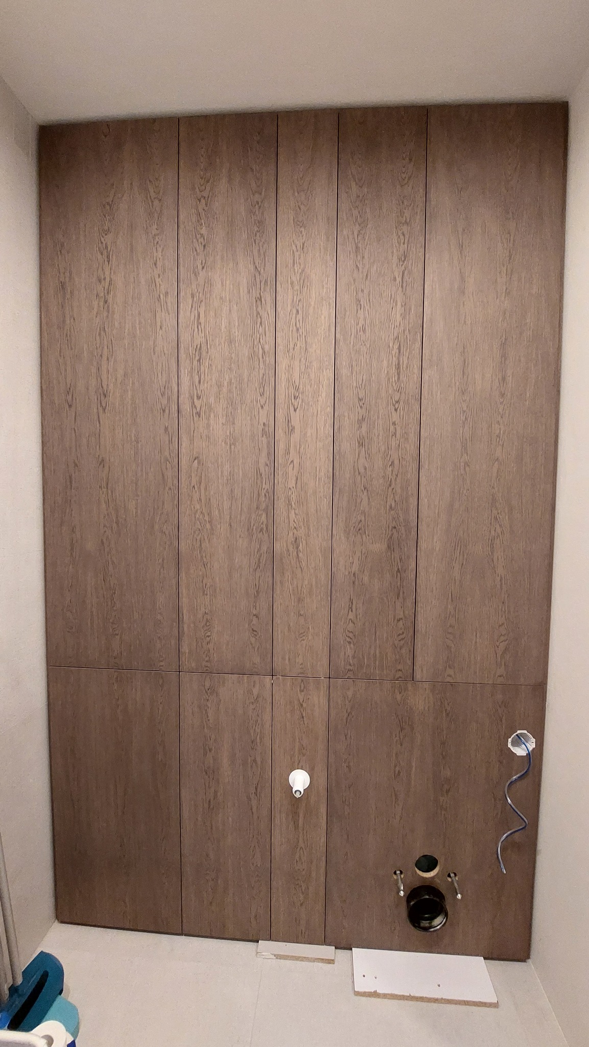 Wysoka zabudowa z drewnopodobnych paneli w łazience, z widocznymi przyłączami sanitarnymi i elektrycznymi. Ujęcie od dołu, podkreślające pionowy układ paneli.