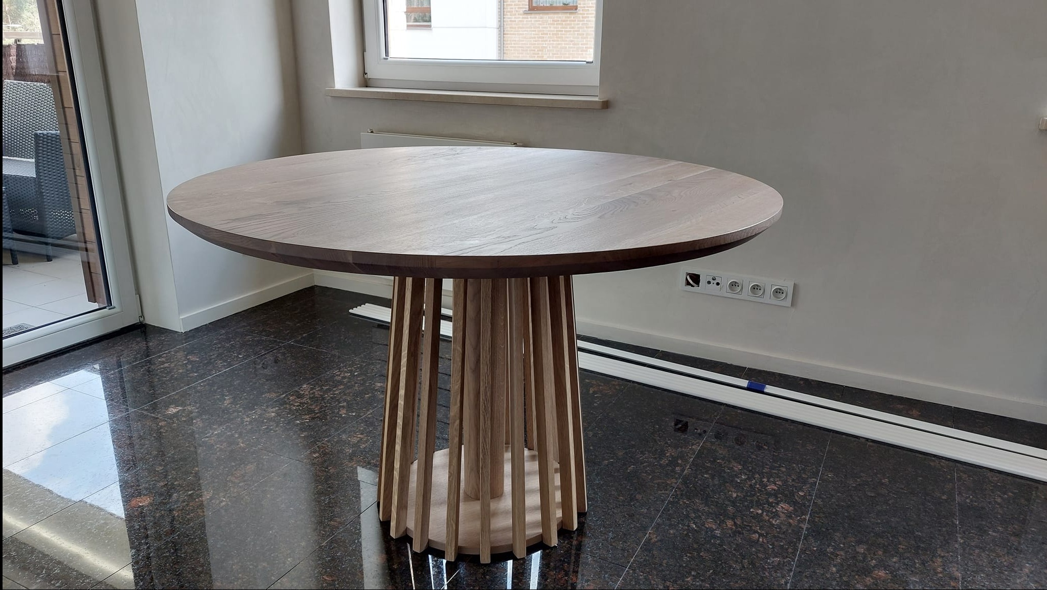 Okrągły stół z blatem z jasnego drewna i unikalną podstawą z pionowych listewek, stojący na ciemnej, błyszczącej podłodze w jasnym pomieszczeniu.