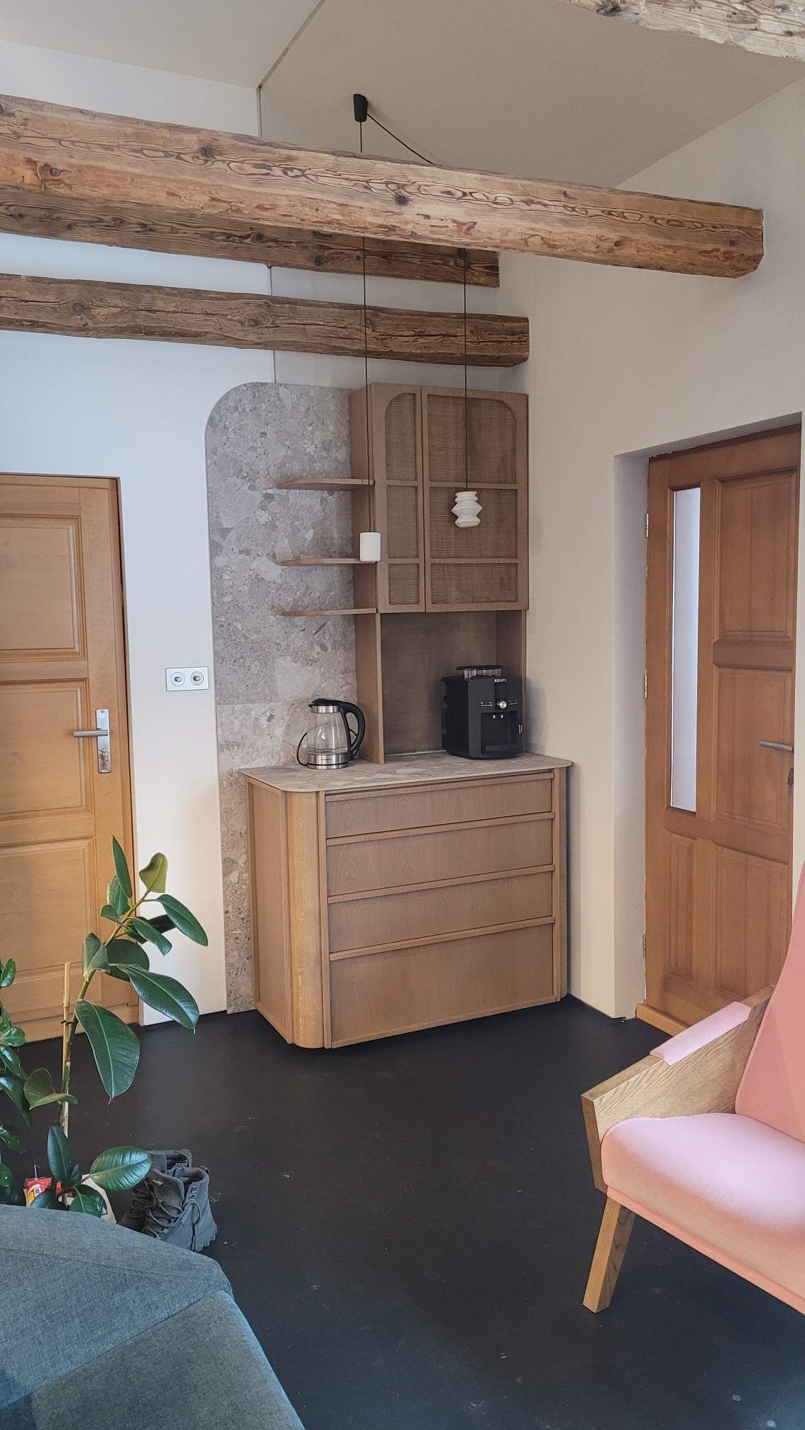 Drewniana zabudowa wnęki z komodą, szafką i półkami, uzupełniona o sprzęt AGD. Widoczne drewniane belki stropowe i drzwi. Całość w neutralnej kolorystyce.