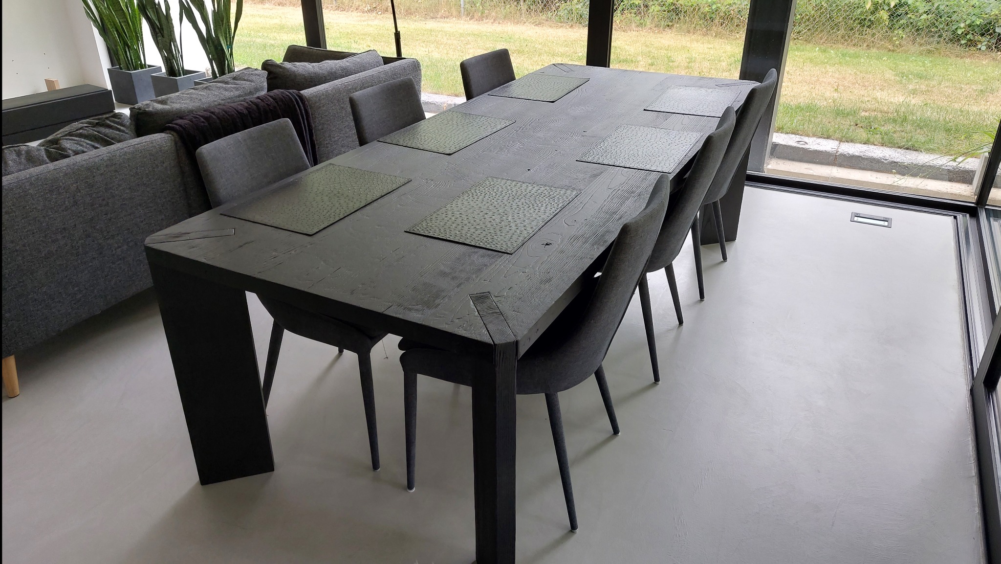 Stół z ciemnego drewna z sześcioma krzesłami w nowoczesnym wnętrzu z dużym oknem. Na stole leżą podkładki. Widoczny fragment sofy w tle.