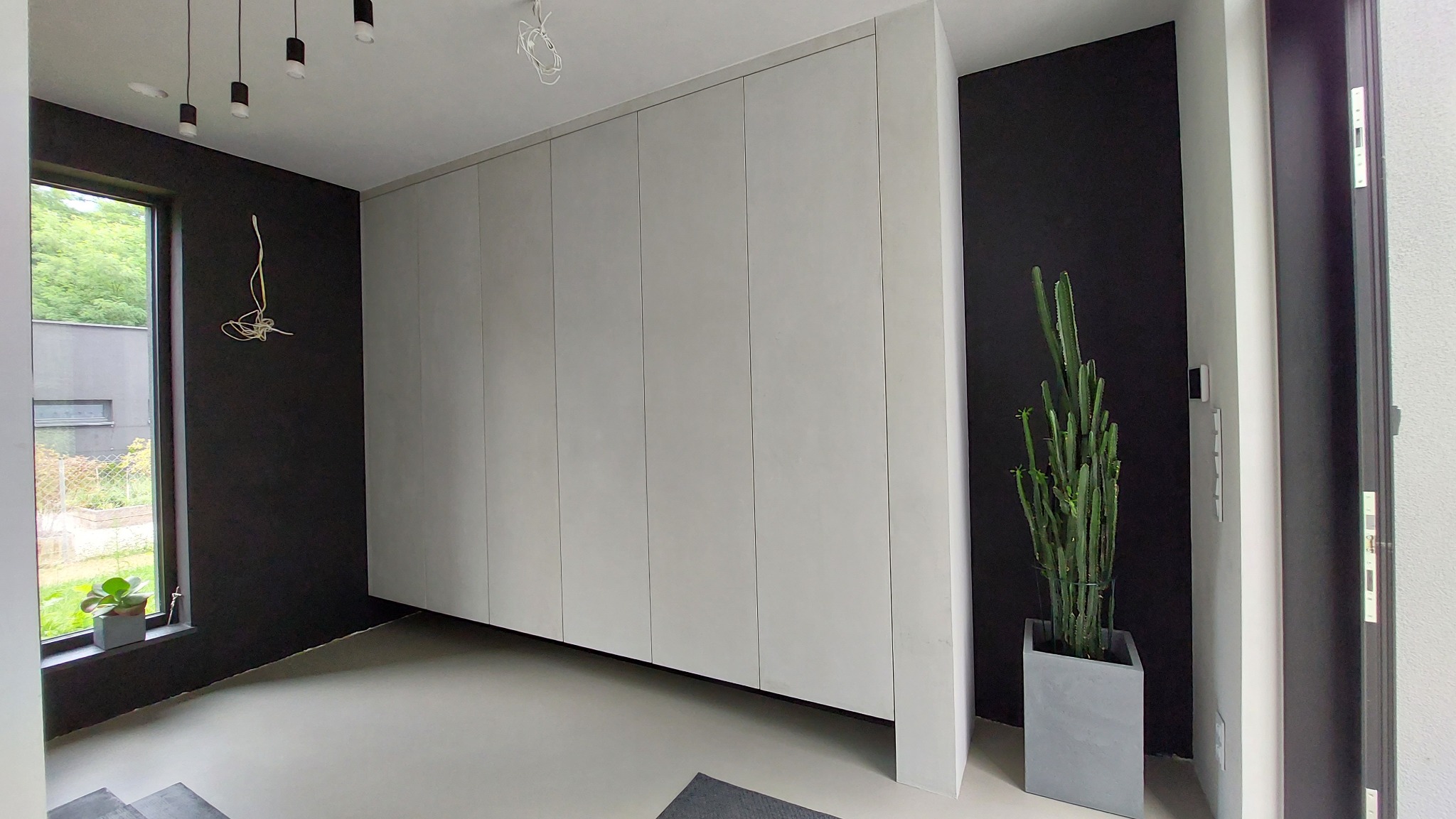 Minimalistyczna szafa wnękowa z jasnymi frontami, wkomponowana w nowoczesne wnętrze z czarnymi ścianami i betonową podłogą, obok drzwi i kaktusa w donicy.