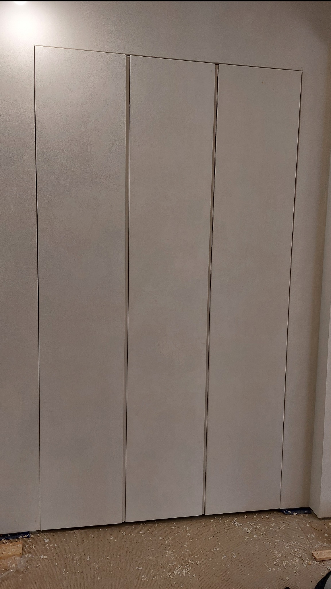 Jasna, wbudowana szafa z trzema drzwiami, zlicowana ze ścianą. Na podłodze widoczne wióry, świadczące o świeżym montażu. Minimalistyczny design.