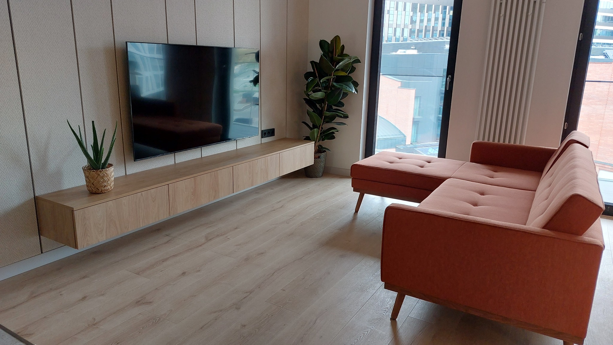 Nowoczesny salon z drewnianą szafką RTV pod telewizorem,  roślinami doniczkowymi i narożnikiem w odcieniach brzoskwini. Widok na miasto z okna.