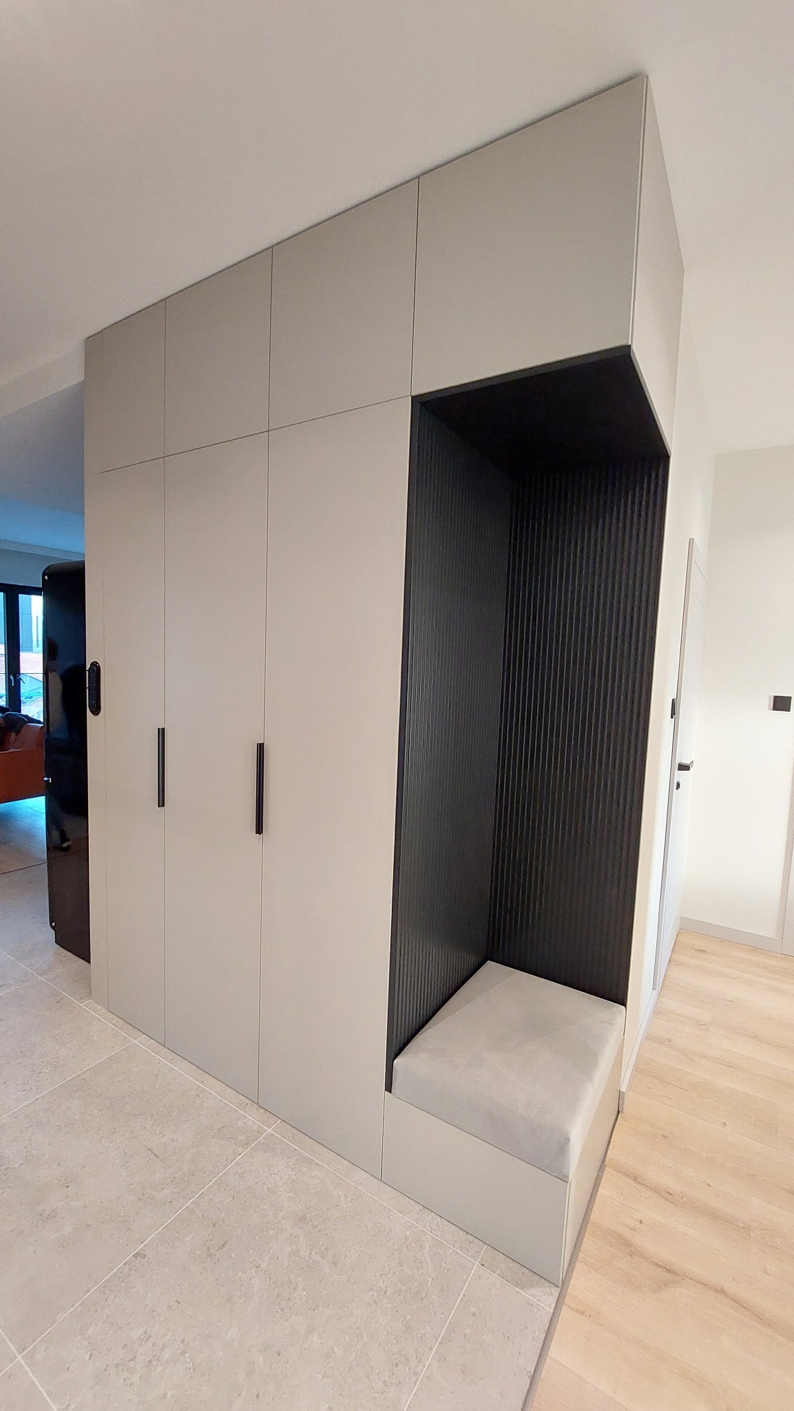 Szara szafa wnękowa z siedziskiem w przedpokoju. Minimalistyczny design, czarne, pionowe lamele w otwartej wnęce. Jasna podłoga z płytek i drewna.