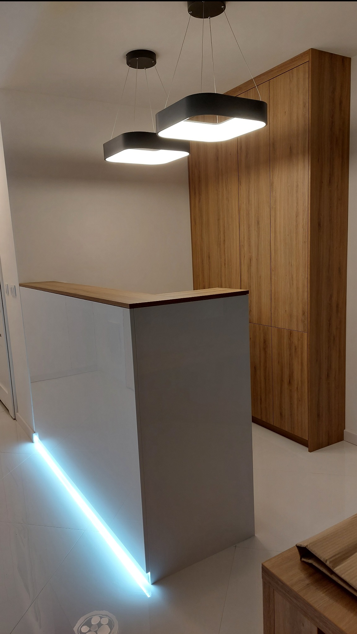 Biała, podświetlana lada z drewnianym blatem i wysoka szafa z drewnopodobnymi frontami. Dwie nowoczesne lampy sufitowe. Minimalistyczne wnętrze.
