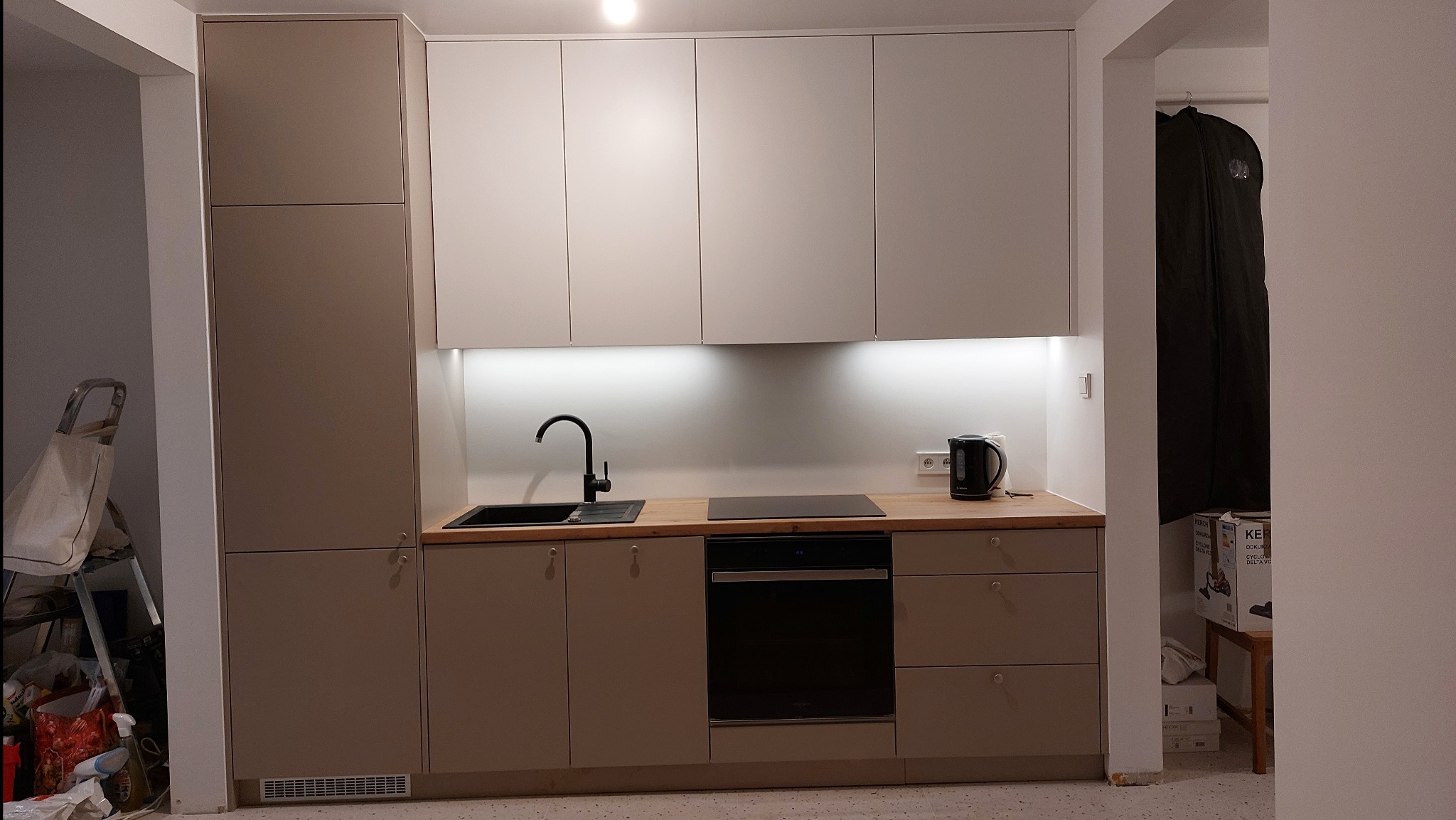 Nowoczesna kuchnia z beżowo-białymi szafkami, drewnianym blatem i czarną armaturą. Podświetlenie LED nad blatem. W tle widoczny fragment innego pomieszczenia.