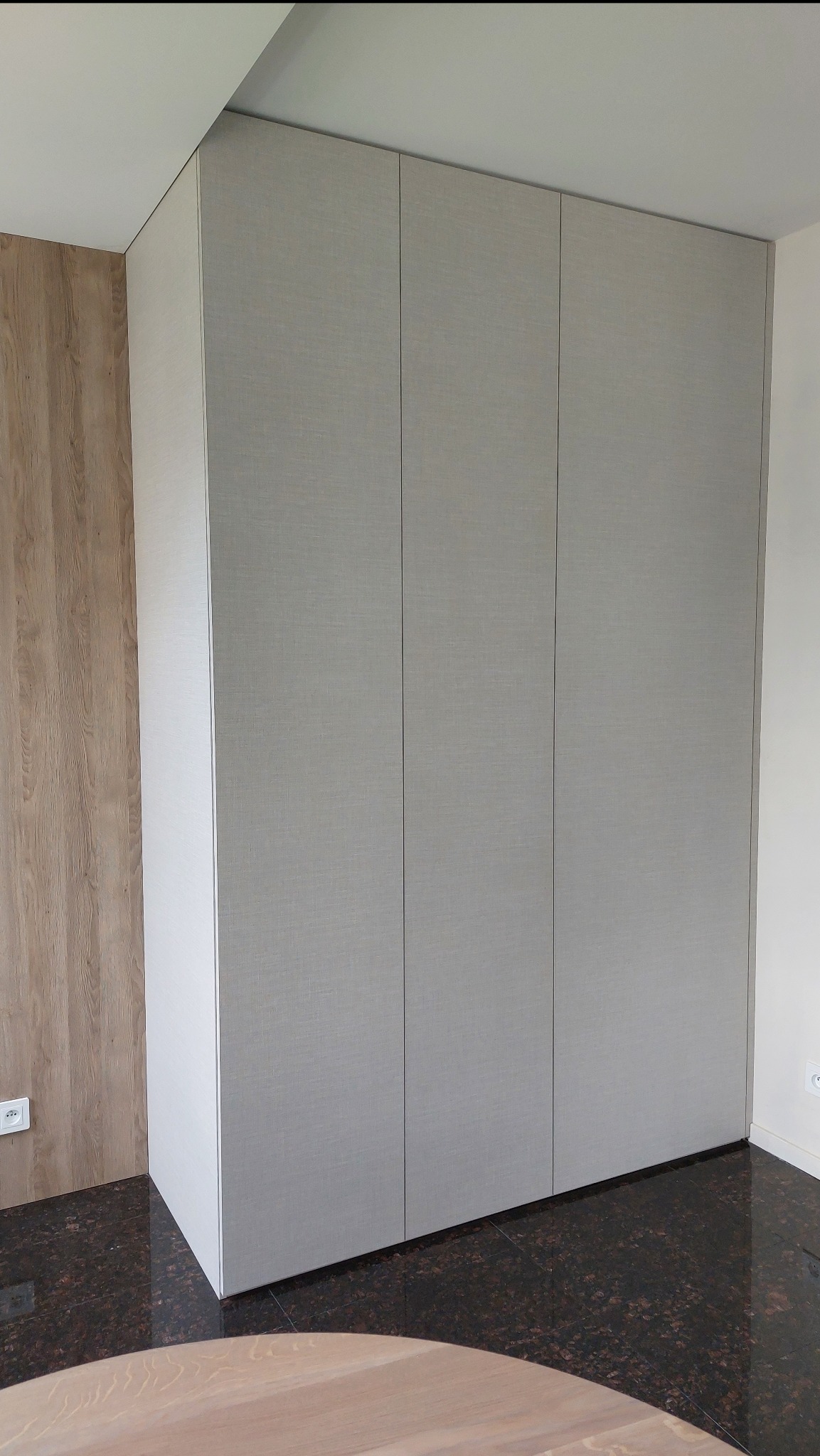 Jasnoszara szafa wnękowa z trzema drzwiami, dopasowana do sufitu, na tle ściany z drewnianą okładziną i ciemnej podłogi. Widoczny fragment okrągłego blatu stołu.