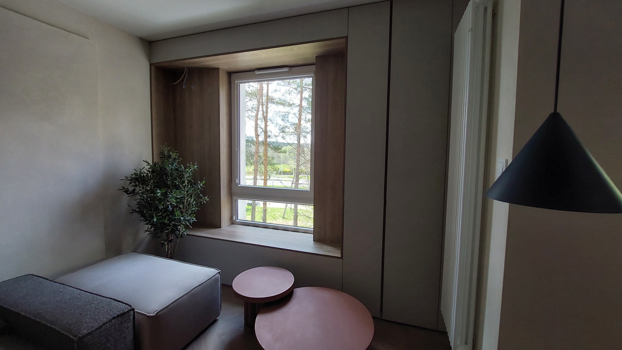 Nowoczesny pokój z oknem w drewnianej zabudowie, w tle widok na zieleń. Minimalistyczne meble i oświetlenie tworzą przytulną atmosferę.