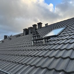 OL-DACH DEKARSTWO - Grafitowy dach z dachówki ceramicznej, z oknem dachowym i kominami na tle pochmurnego nieba. Widoczne zabezpieczenia przeciwśniegowe.