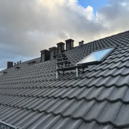 Grafitowy dach z dachówki ceramicznej, z oknem dachowym i kominami na tle pochmurnego nieba. Widoczne zabezpieczenia przeciwśniegowe.