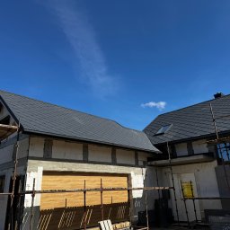 OL-DACH DEKARSTWO - Dom w trakcie budowy z szarym dachem z dachówki, widoczne rusztowania i błękitne niebo. Dach pokryty dachówką, okna wstawione, brama garażowa zamontowana.