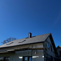 OL-DACH DEKARSTWO - Dom w trakcie krycia dachu szarą dachówką z widocznymi oknami dachowymi i kominem. Częściowo otynkowana elewacja i rusztowanie. Błękitne niebo w tle.