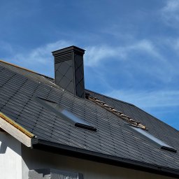 OL-DACH DEKARSTWO - Czarny dach z dachówki ceramicznej z oknami dachowymi i kominem na tle błękitnego nieba. Widoczne elementy konstrukcji dachu i rynna.