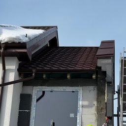 OL-DACH DEKARSTWO - Montaż brązowej blachodachówki trapezowej przy oknie. Widoczny dekarz w żółtej kurtce uszczelniający pianką przestrzeń. Część dachu pokryta śniegiem.