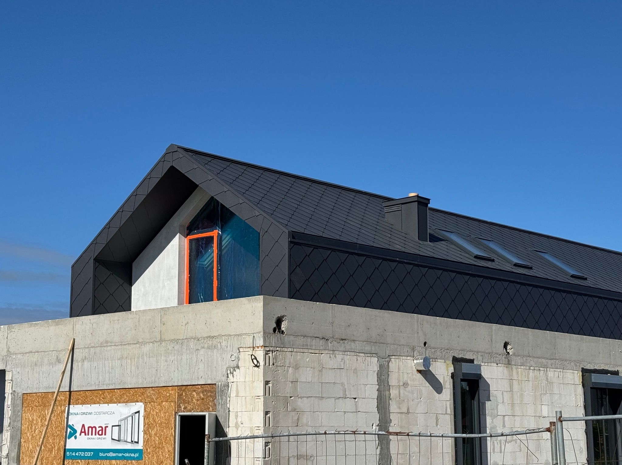 Nowoczesny dom w trakcie budowy z grafitowym dachem z dachówki karpiówki i oknami dachowymi, widok z dołu na tle błękitnego nieba.