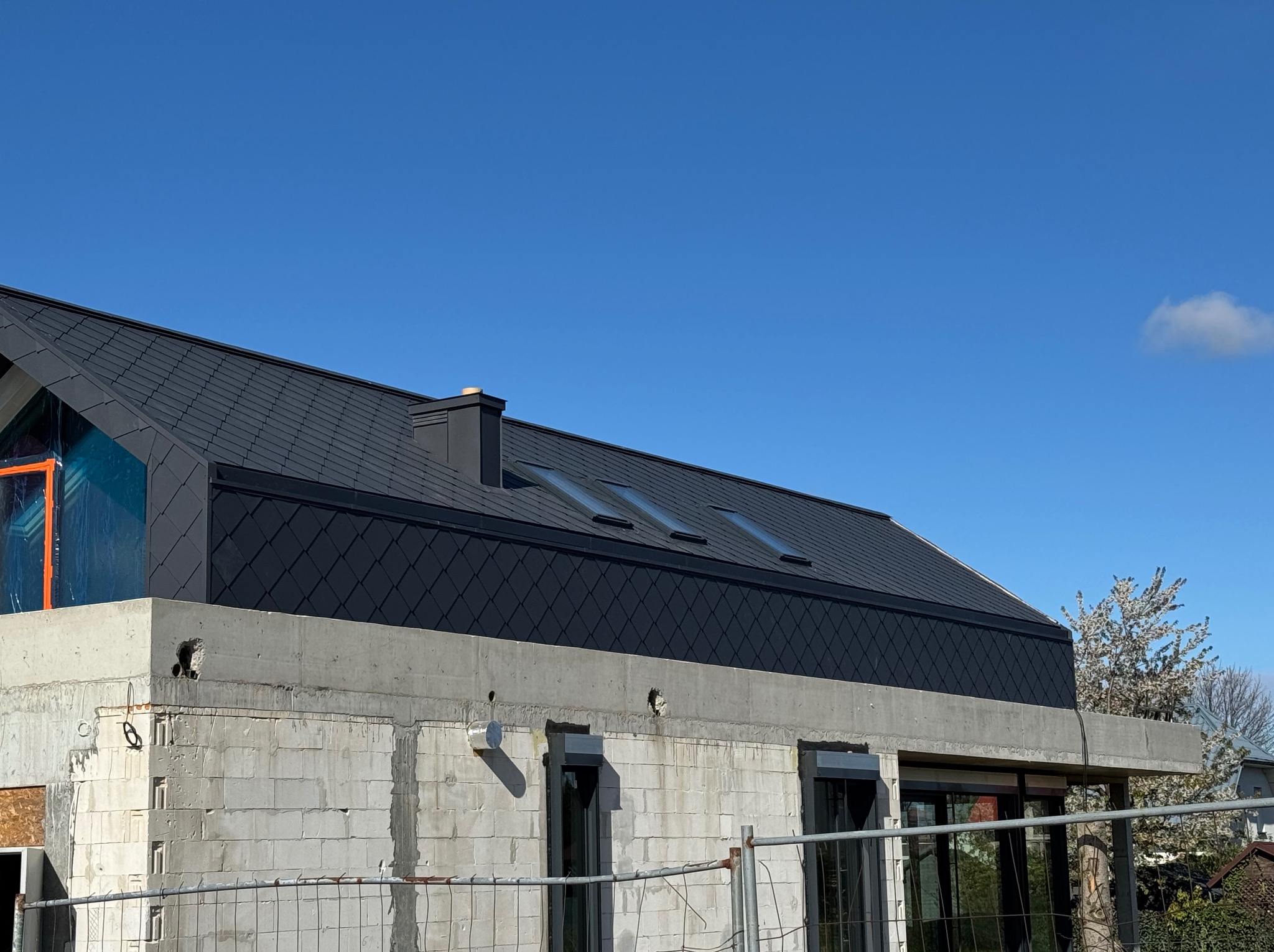 Nowoczesny dom w trakcie budowy, z ciemnym dachem krytym romboidalną dachówką i oknami dachowymi. Widoczne elementy konstrukcyjne i zabezpieczenia.