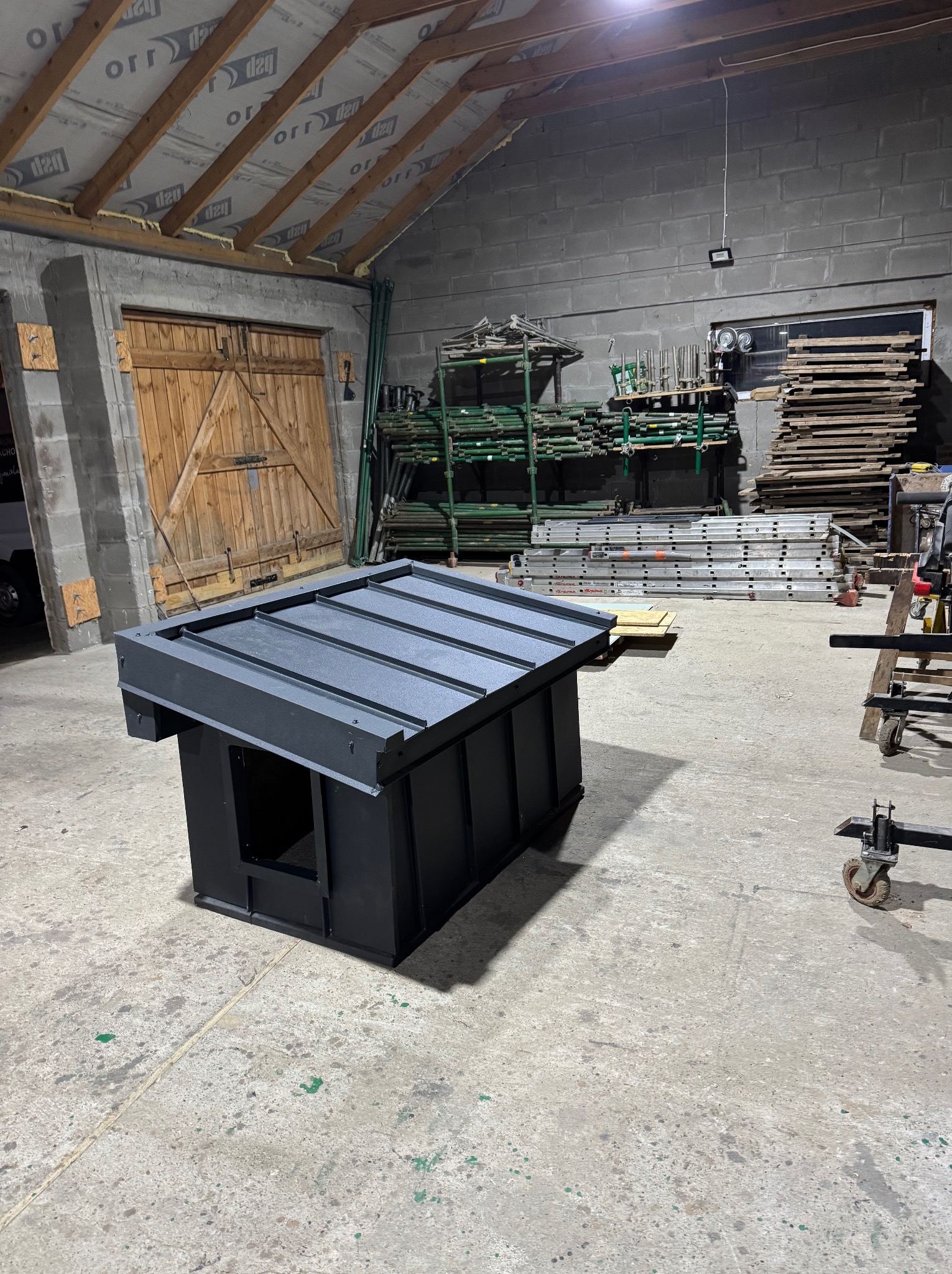 Gotowy element dachu w warsztacie: czarny, modułowy fragment z blachy, przygotowany do montażu. W tle rusztowania i drewno.