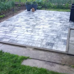 Ułożona kostka brukowa w szaro-białej kolorystyce, na tle zielonego ogrodu z roślinami w donicach. Widoczne elementy małej architektury ogrodowej.