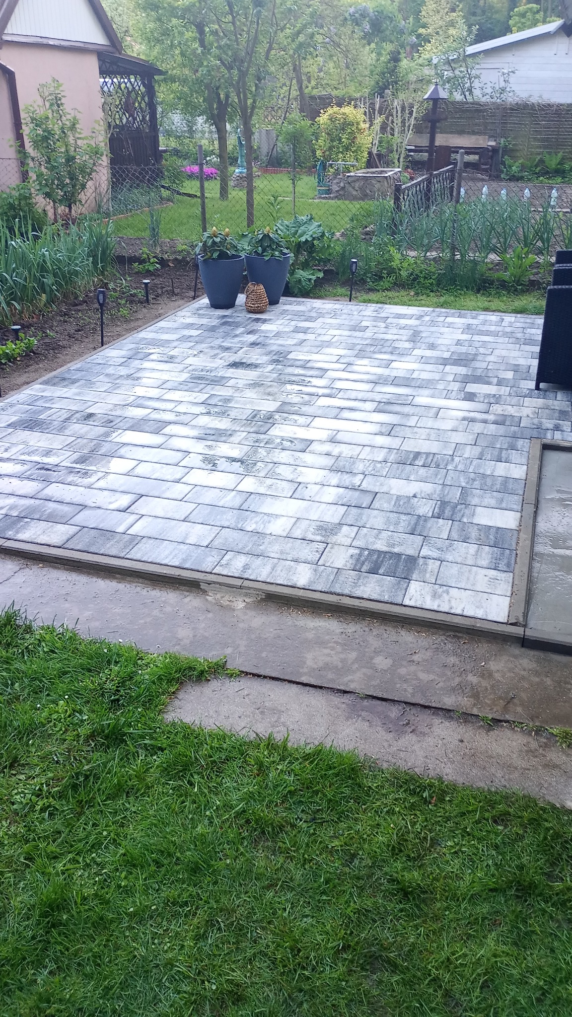 Ułożona kostka brukowa w szaro-białej kolorystyce, na tle zielonego ogrodu z roślinami w donicach. Widoczne elementy małej architektury ogrodowej.