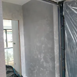 Szara ściana z efektem dekoracyjnym w nowym wnętrzu, widoczny fragment futryny drzwiowej z zabezpieczeniem folią. Podłoga wyłożona białym gresem.