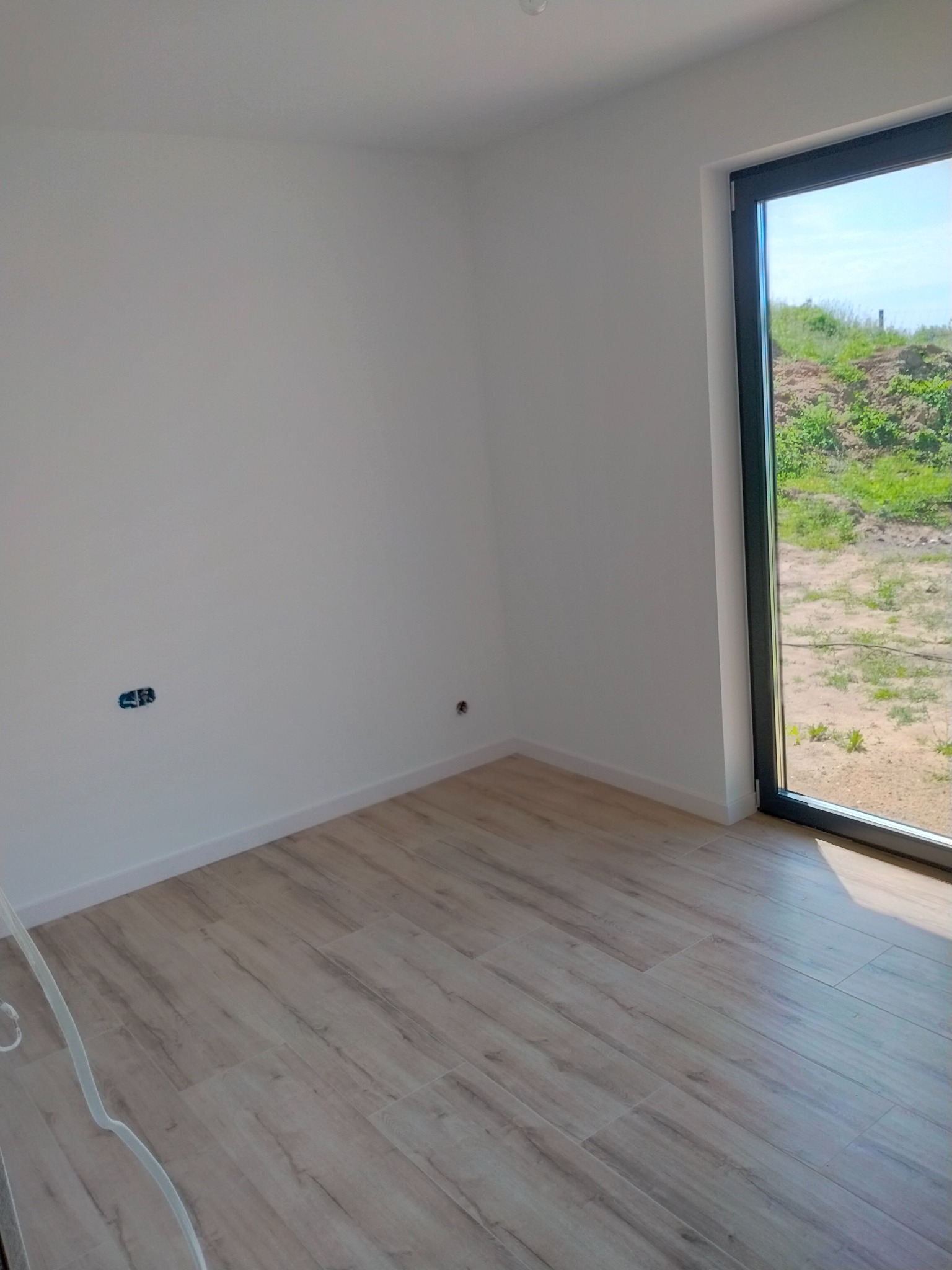 Jasne panele podłogowe w pokoju z białymi ścianami i dużym oknem z widokiem na zieleń. Nowe gniazdka elektryczne. Minimalistyczne wykończenie.