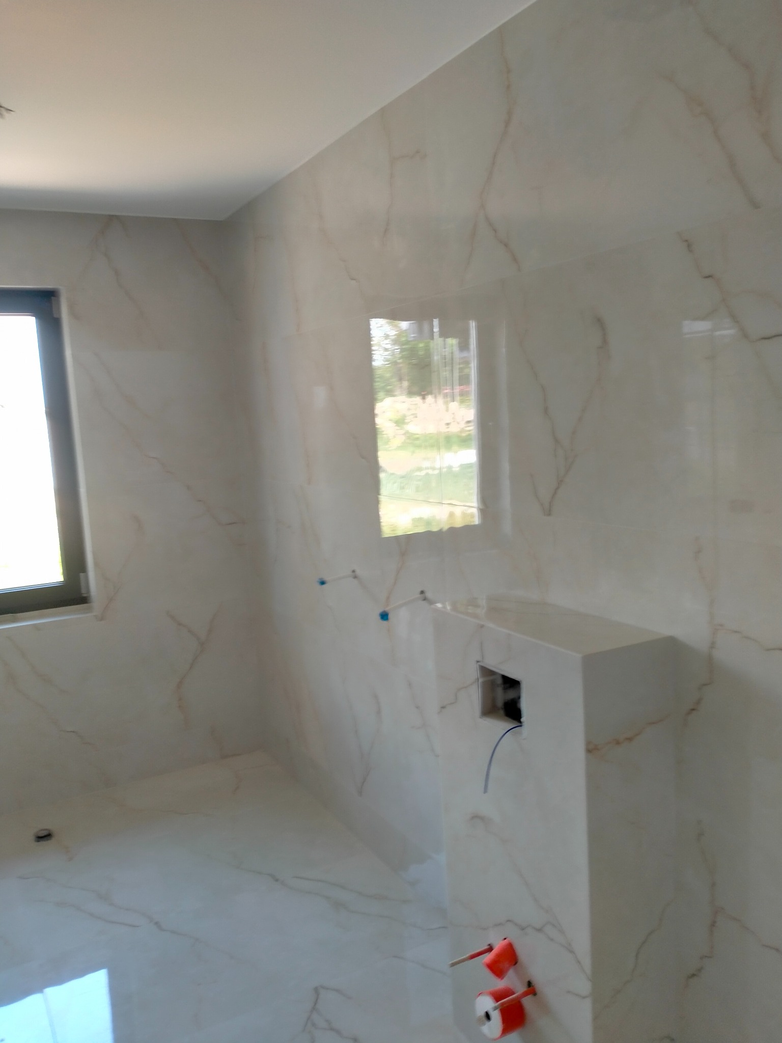 Jasna łazienka z marmurowymi płytkami, widoczne okno i zabudowa podtynkowego WC z wystającymi elementami instalacji. Minimalistyczny design, neutralna kolorystyka.