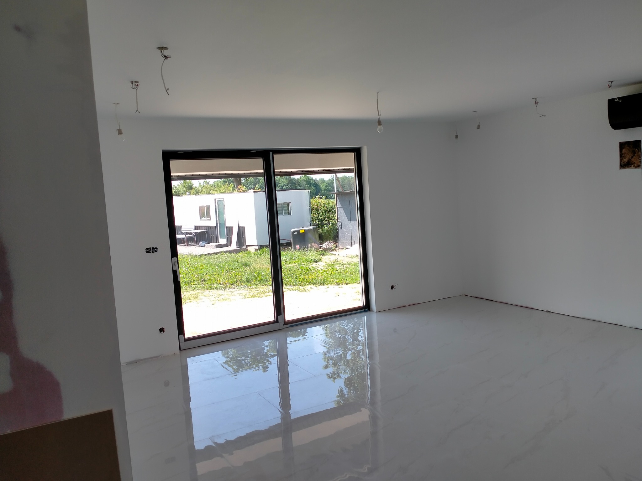 Jasne wnętrze po remoncie z białymi ścianami, podłogą z płytek i dużym oknem przesuwnym. Widoczne niedokończone podłączenia oświetlenia.