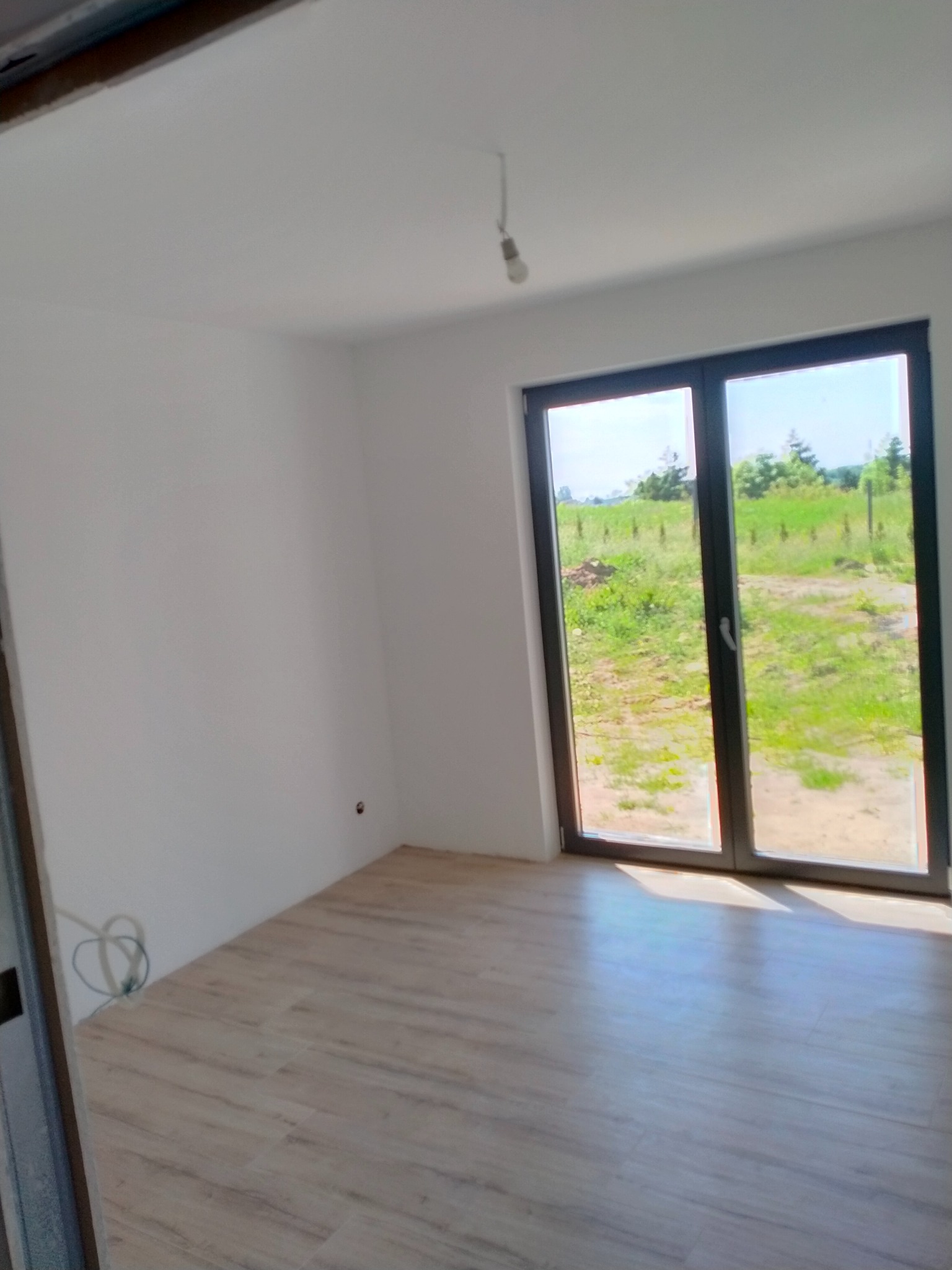 Jasne panele podłogowe w nowym, pustym pokoju z białymi ścianami i dużym oknem z widokiem na zieleń. Kabel wystający ze ściany.