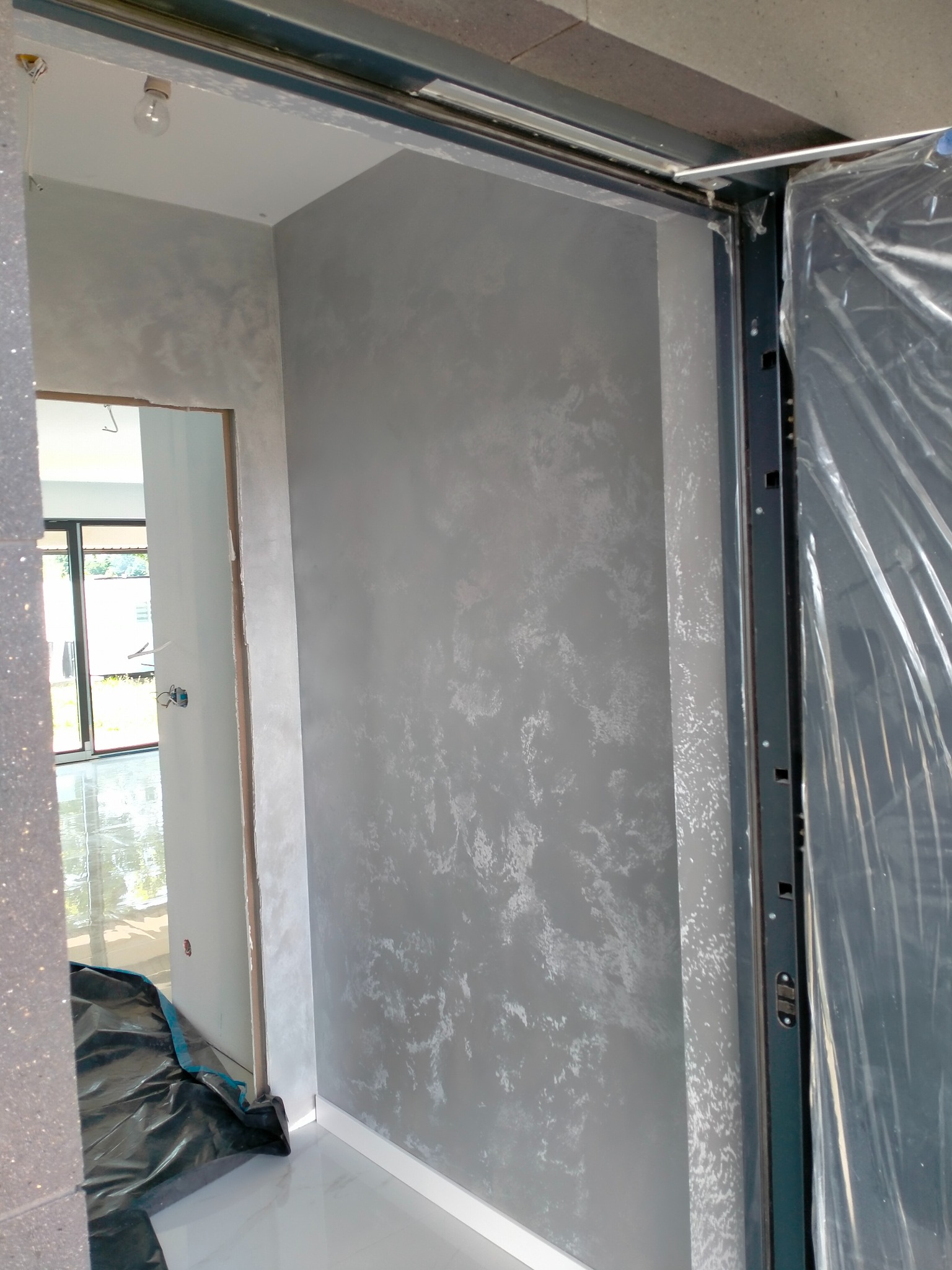 Szara ściana z efektem dekoracyjnym w nowym wnętrzu, widoczny fragment futryny drzwiowej z zabezpieczeniem folią. Podłoga wyłożona białym gresem.