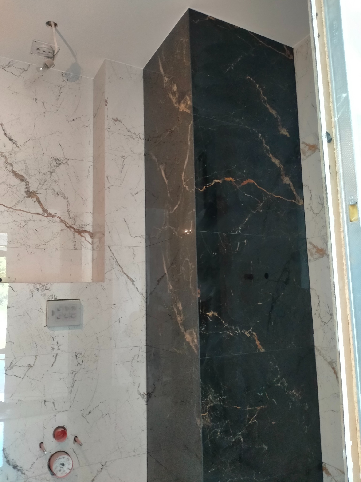 Wykończona ściana w łazience z marmurowymi płytkami w odcieniach bieli i czerni, z widoczną instalacją sanitarną i włącznikiem. Detal wykończenia wnętrza.