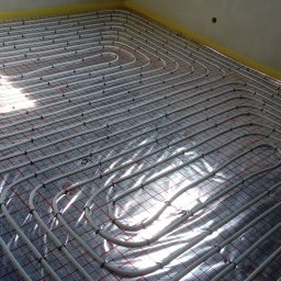 PRZEDSIĘBIORSTWO INSTALACYJNE SOTMET SPÓŁKA Z OGRANICZONĄ ODPOWIEDZIALNOŚCIĄ - Ułożone białe rury ogrzewania podłogowego na folii z czerwoną kratką, mocowane czarnymi uchwytami. Widoczne odbicia światła. Żółta taśma przy ścianie.