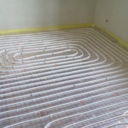 PRZEDSIĘBIORSTWO INSTALACYJNE SOTMET SPÓŁKA Z OGRANICZONĄ ODPOWIEDZIALNOŚCIĄ - Białe rury ogrzewania podłogowego ułożone na folii z czerwoną siatką w jasnym pomieszczeniu, zabezpieczone żółtą taśmą przy ścianach. Widok z góry na instalację.